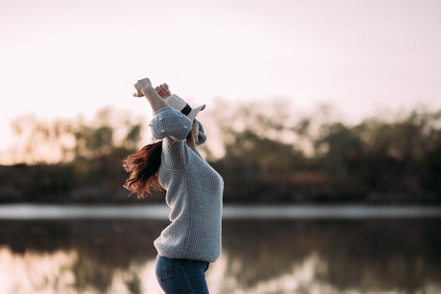 Femme brune dansant joyeusement au bord d'une rivière avec un chapeau levant les bras