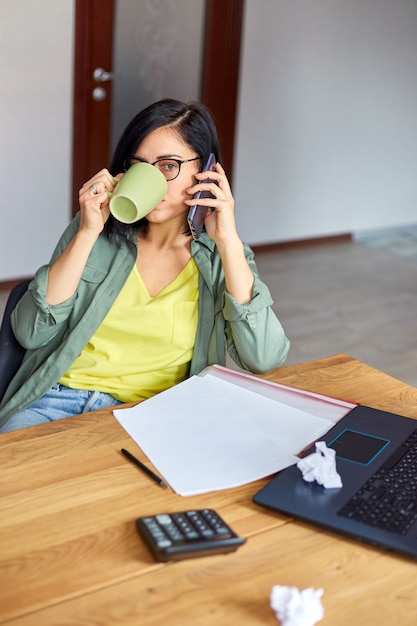 Femme brune élégante dans des verres assis à une table en bois avec bloc-notes et ayant un appel téléphonique