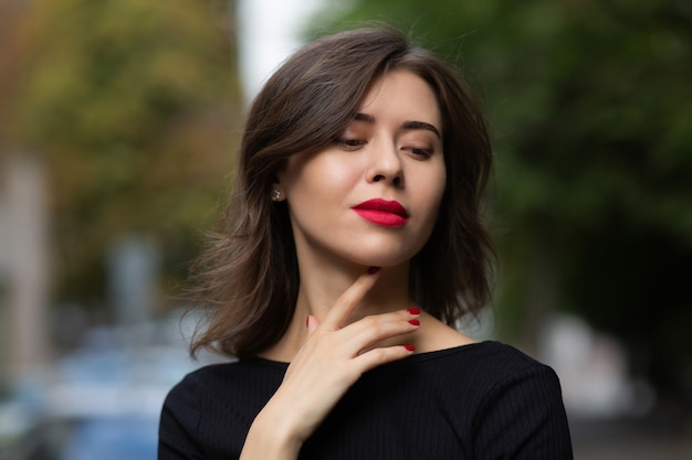 Une femme brune élégante et sensuelle aux lèvres rouges porte une robe noire posant dans la rue. Photo en gros plan