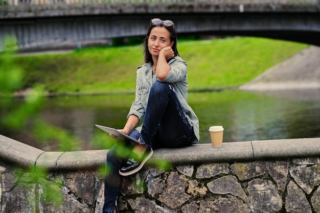Une femme brune est assise sur un pont et tient une tablette PC.