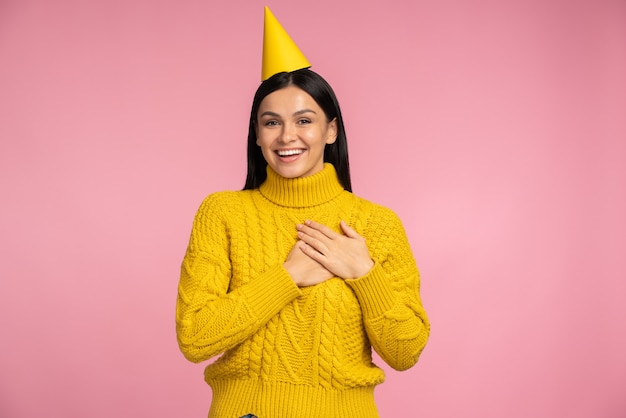 Femme brune extrêmement heureuse en chapeau de fête appréciant sincèrement, recevant des félicitations et des cadeaux. Studio intérieur tourné isolé sur fond rose