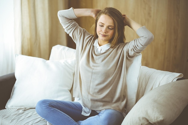 Une femme brune heureuse met les mains derrière la tête assise appuyée sur le canapé une femme européenne profite d'un week-end paresseux ou de vacances dans un salon confortable, la femme au foyer se détend se sent satisfaite après les travaux ménagers.