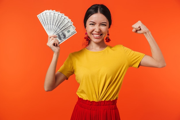 femme brune hispanique des années 20 vêtue d'une jupe souriante tout en tenant un tas de billets d'argent isolés sur un mur rouge