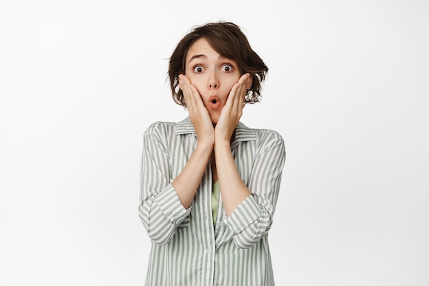 Une femme brune intriguée et surprise dit wow, se tient la main sur le visage, a l'air étonnée, réagit à des nouvelles géniales, debout en chemise sur fond blanc