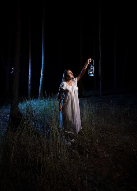 Femme brune avec une lanterne à gaz éclairant la forêt la nuit