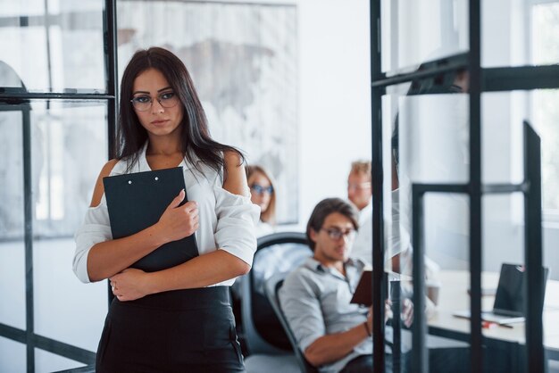 Une femme brune à lunettes avec un bloc-notes dans les mains se tient dans le bureau en face des employés.