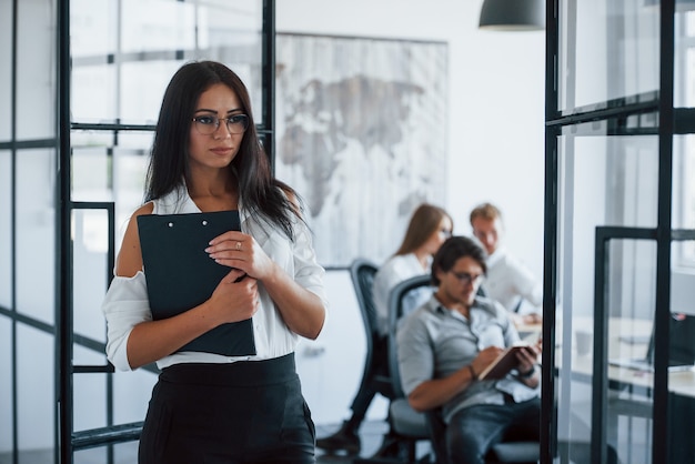 Une femme brune à lunettes avec un bloc-notes dans les mains se tient dans le bureau en face des employés.