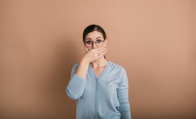 Une femme brune avec des lunettes dans un pull bleu se tient debout et se couvrit la bouche de ses mains sur un fond beige avec une place pour le texte