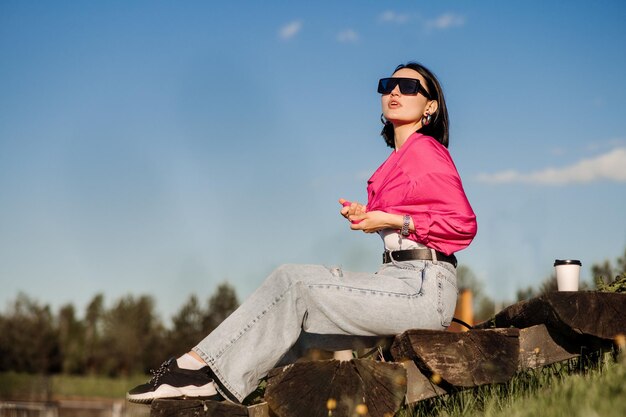 Femme brune à lunettes de soleil et chemise rose assise et relaxante dans le parc sur fond de ciel bleu