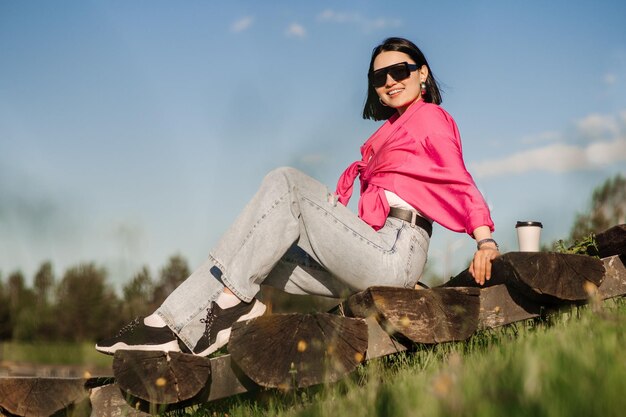 Femme brune à lunettes de soleil et chemise rose assise et relaxante dans le parc sur fond de ciel bleu