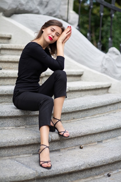 Une femme brune luxueuse aux lèvres rouges porte un costume noir assis sur les escaliers en béton gris