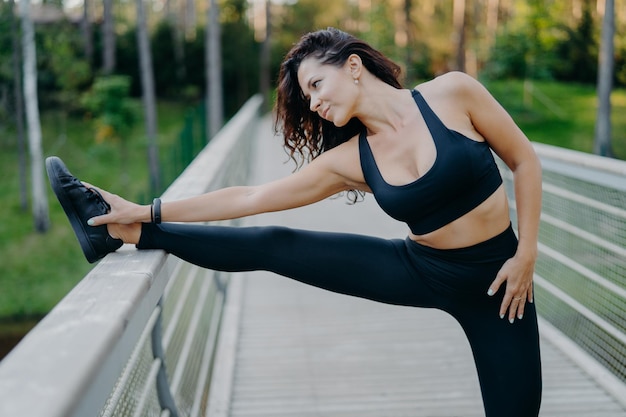 Une femme brune mince et sportive vêtue d'un haut court et de leggings étire les jambes sur le pont se réchauffe avant la pose de la course du matin en plein air veut avoir un corps mince et une bonne santé