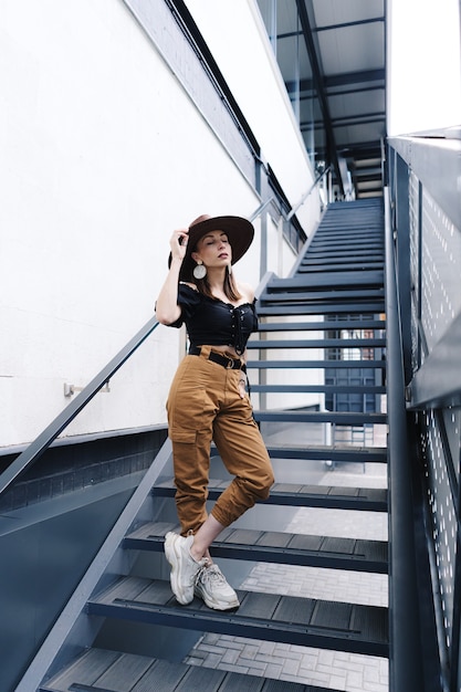 Femme brune à la mode aux cheveux longs, portant un grand chapeau en osier élégant, posant dans les escaliers.