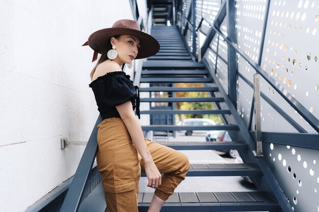 Femme brune à la mode aux cheveux longs, portant un grand chapeau en osier élégant, posant dans les escaliers.