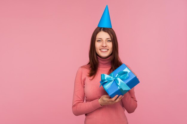 Femme brune positive optimiste en chapeau conique sur la tête tenant un cadeau avec un arc et regardant la caméra avec un sourire à pleines dents, des cadeaux et des bonus, célébration. Studio intérieur tourné isolé sur fond rose