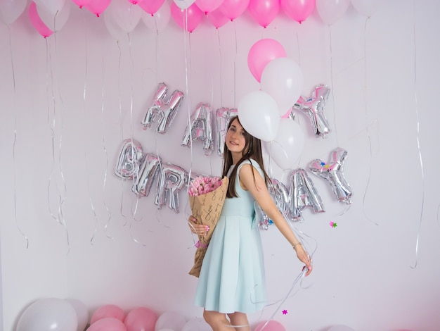 Femme brune en robe menthe tenant un bouquet de tulipes. Décorations d'anniversaire avec ballons de couleur blanche et rose, bouquet de tulipes et confettis pour la fête sur un mur blanc. Joyeux anniversaire anniversaire.