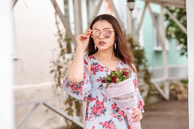 Femme brune romantique en lunettes de soleil miroir et robe bleu clair avec bouquet de pivoines dans ses mains.