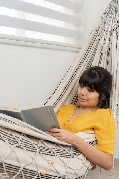 Une femme brune se trouve dans un hamac à l'intérieur, lit un livre et boit du jus de fruit