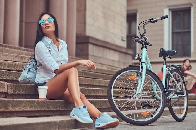 Une femme brune sexy portant un chemisier et un short en jean à lunettes de soleil, se détendant après avoir fait du vélo, assise avec une tasse de café sur les marches d'une ville.