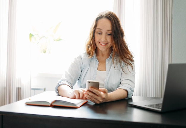 Femme brune souriante adulte faisant des notes dans un livre quotidien et utilisant un téléphone portable avec un ordinateur portable à la maison