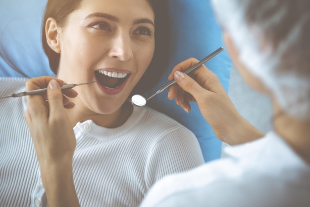 Femme brune souriante en cours d'examen par un dentiste à la clinique dentaire. Mains d'un médecin tenant des instruments dentaires près de la bouche du patient. Dents saines et concept de médecine.