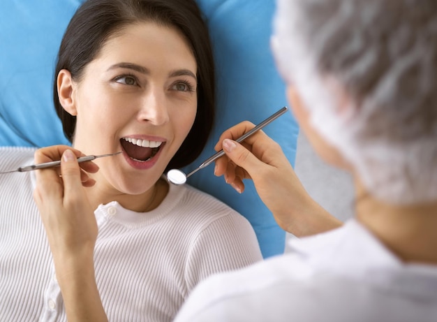 Femme brune souriante en cours d'examen par un dentiste à la clinique dentaire. Mains d'un médecin tenant des instruments dentaires près de la bouche du patient. Dents saines et concept de médecine.