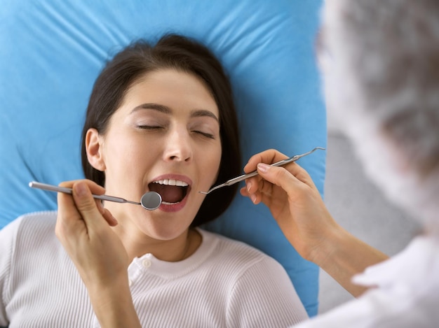 Une femme brune souriante examinée par un dentiste dans une clinique dentaire. Les mains d'un médecin tenant des instruments dentaires près de la bouche du patient. Des dents saines et un concept médical.