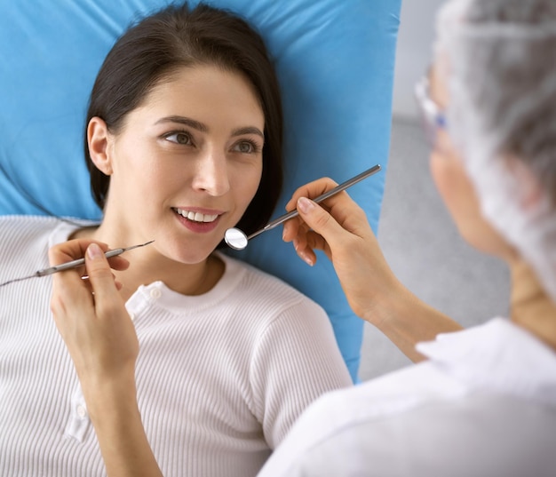 Une femme brune souriante examinée par un dentiste dans une clinique dentaire. Les mains d'un médecin tenant des instruments dentaires près de la bouche du patient. Des dents saines et un concept médical.