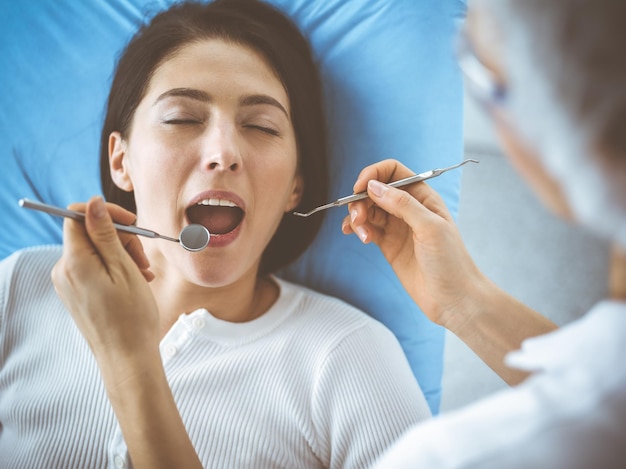 Une femme brune souriante examinée par un dentiste dans une clinique dentaire. Les mains d'un médecin tenant des instruments dentaires près de la bouche du patient. Des dents saines et un concept médical.