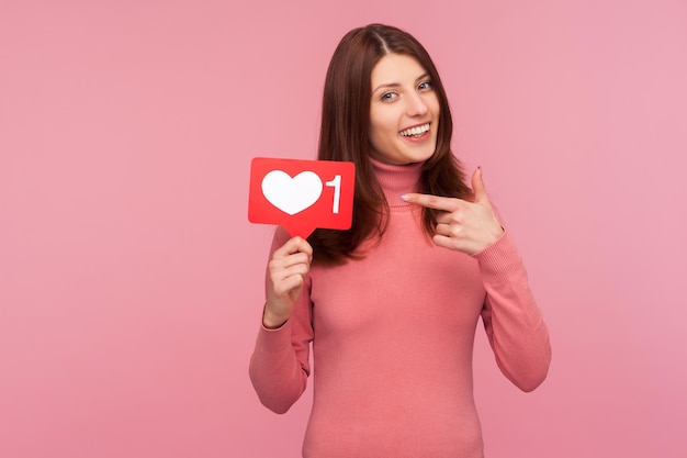 Femme brune souriante en pull rose pointant du doigt le signe avec comme compteur, blogueur attendant des commentaires et un taux positif. Studio intérieur tourné isolé sur fond rose