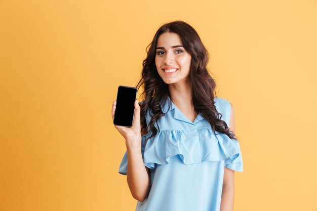 Femme brune souriante en robe bleue montrant un écran de smartphone vierge isolé