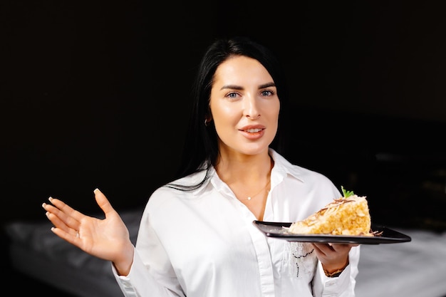 Femme brune souriante tenant une assiette avec du gâteau dans la chambre de son appartement