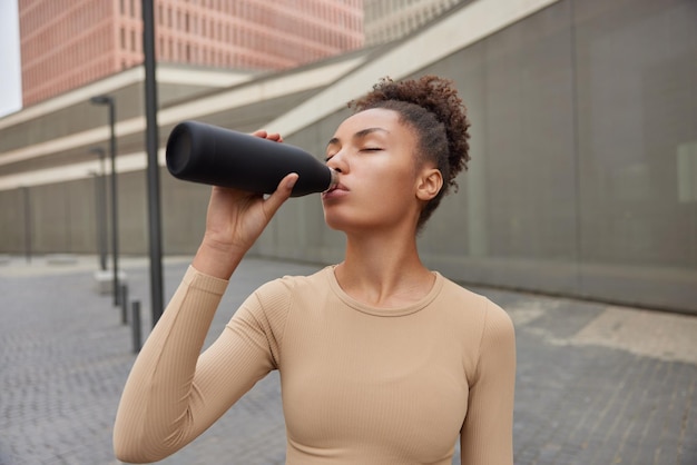 Une femme brune sportive active a soif après l'entraînement boit de l'eau fraîche de la bouteille fatiguée après l'entraînement vêtue de vêtements de sport pose contre un cadre urbain flou Concept de sport et de santé