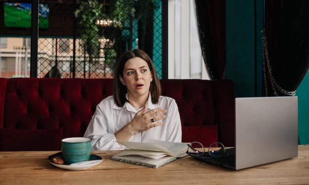 Une femme brune surprise dans un chemisier blanc est assise à une table avec un ordinateur portable et un ordinateur portable dans un café