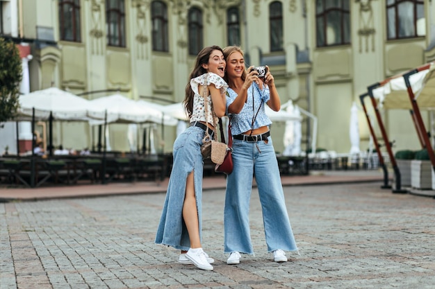 Femme brune surprise en pantalon denim élégant et chemisier à fleurs blanc pose avec son amie à l'extérieur Dame blonde en jeans et haut bleu prend des photos sur un appareil photo rétro