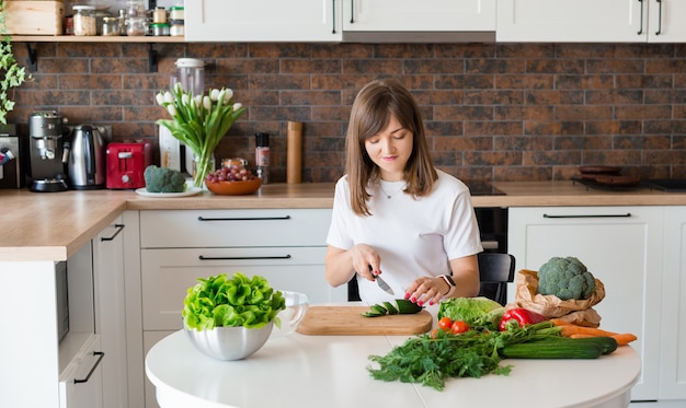 Femme brune en t-shirt blanc préparant une salade de légumes dans la cuisine à la maison, femme coupant des ingrédients sur la table. Nourriture saine, salade végétalienne. Concept de régime.