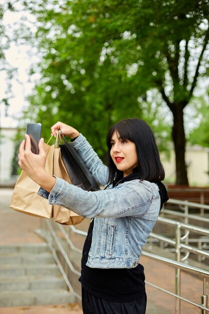 Femme brune tenant des sacs en papier et prenant selfie par smartphone dans le parc, fille après le shopping.