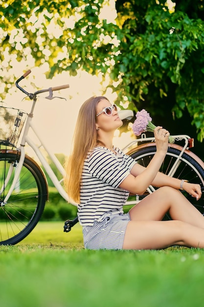 Une femme brune tient un bouquet près d'un vélo de ville dans un parc d'été.