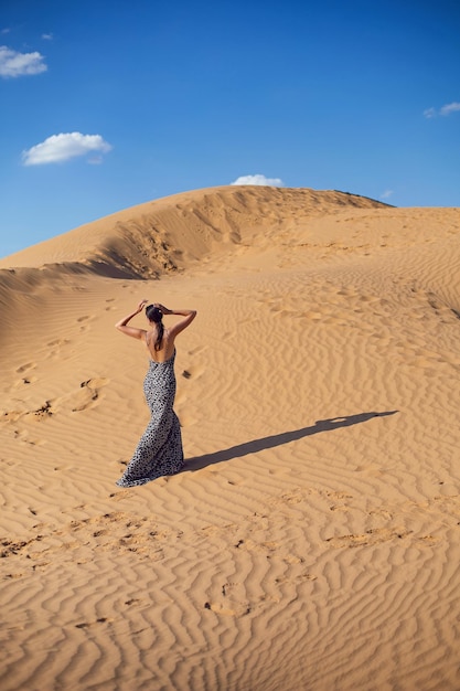Une femme brune vêtue d'une longue robe léopard se tient dos dans le désert au coucher du soleil, allez partout