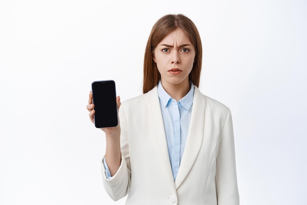 Une femme de bureau de PDG confuse en costume d'affaires fronce les sourcils montre un écran de smartphone vide se plaint et démontre quelque chose de mal sur fond blanc de téléphone