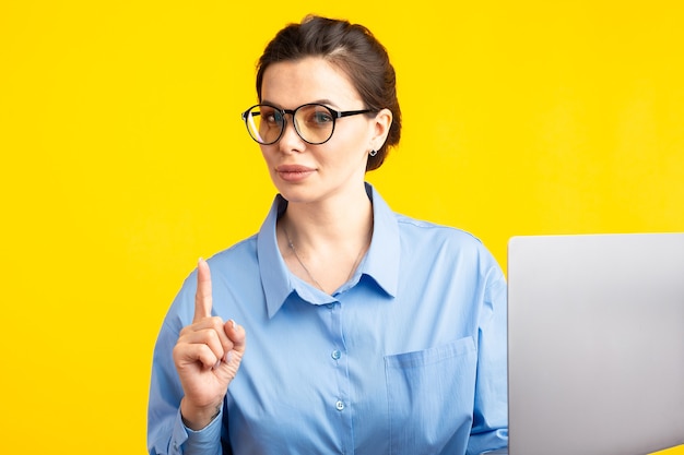 Femme de bureau sérieux dans shurt dans des verres tenant un ordinateur portable et en l'utilisant debout isolé sur fond jaune.