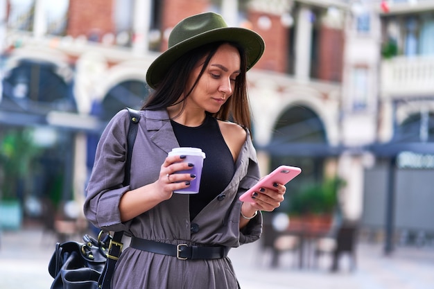 Femme buvant du café et à l'aide de smartphone lors de la marche dans le centre d'une ville européenne