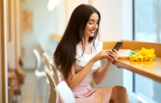 Femme buvant du café et utilisant un téléphone. style de printemps. femme brune au beau printemps. Belle jeune femme avec bouquet de fleurs de tulipes de printemps au café. belle humeur printanière