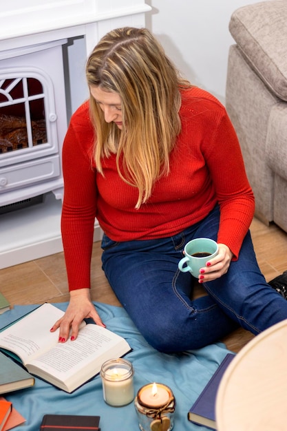 Femme avec café lisant un livre assis sur un sol dans le salon à l'intérieur d'une maison en hiver
