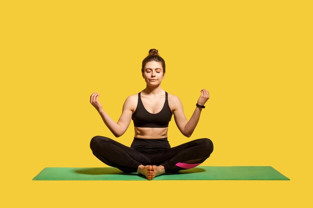 Femme calme concentrée avec un chignon dans des vêtements de sport serrés assis en position du lotus sur un tapis de gym pratiquant le yoga tenant la main dans le geste mudra et méditant en studio intérieur isolé sur jaune