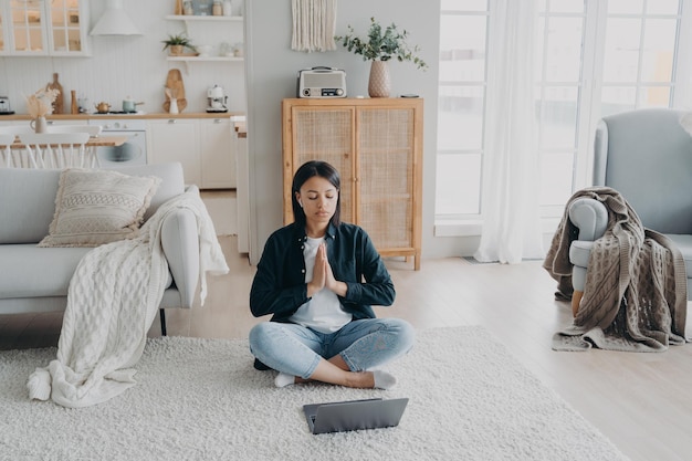 Une femme calme pratique le yoga sur un ordinateur portable a une pause après avoir travaillé assis sur le sol à la maison Soulagement du stress