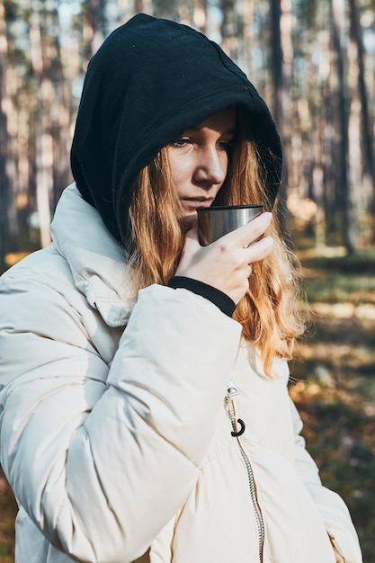 Une femme avec une cape qui fait une pause pendant un voyage d'automne tenant une tasse avec une boisson chaude d'un thermos