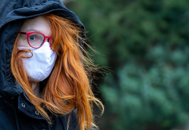 Une femme à capuche aux cheveux roux se tient seule et triste dans un parc. Elle porte un masque respiratoire.