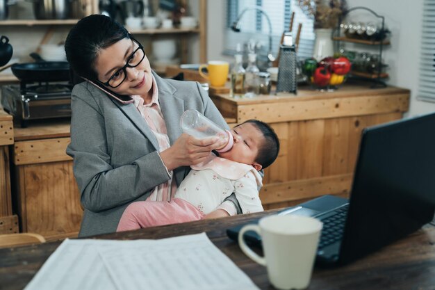 une femme de carrière japonaise occupée inclinant la tête parle avec un smartphone et nourrit bébé. une femme d'affaires asiatique occupée s'occupe du travail au téléphone et traite un enfant dans un bureau à domicile.