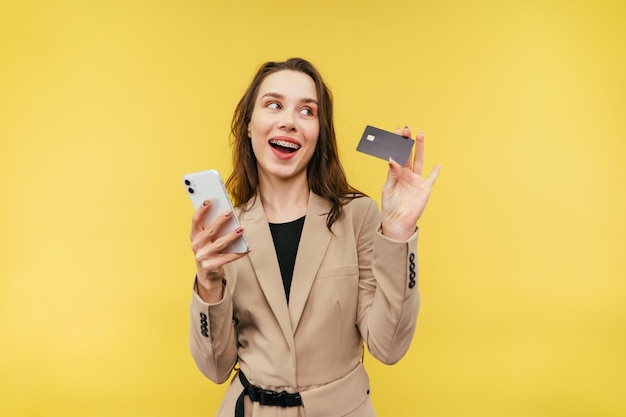 femme avec une carte bancaire en main fait des achats en ligne à partir d'un smartphone sur fond jaune